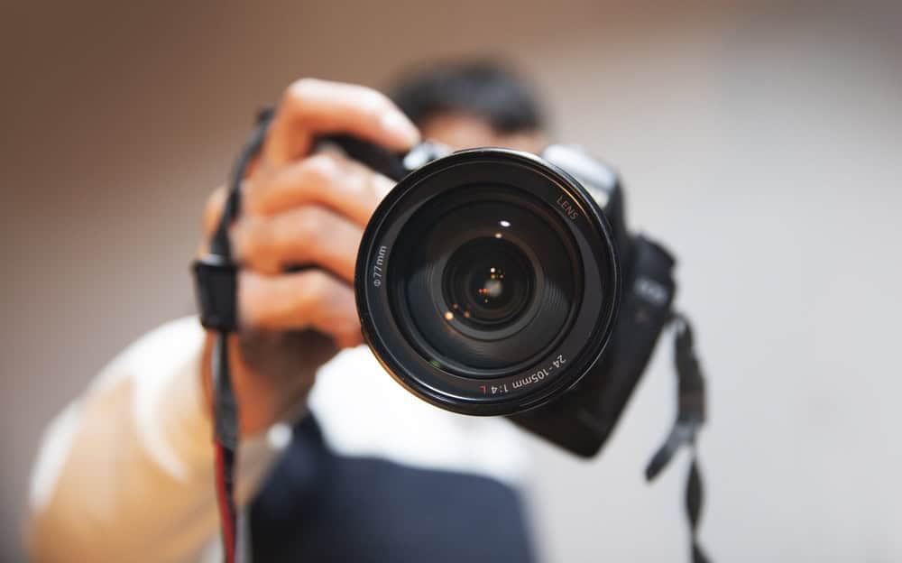 A person holds a DSLR camera close to the lens, partially obscuring their face. The camera is in sharp focus, showing details of the lens, while the background and person are blurred.