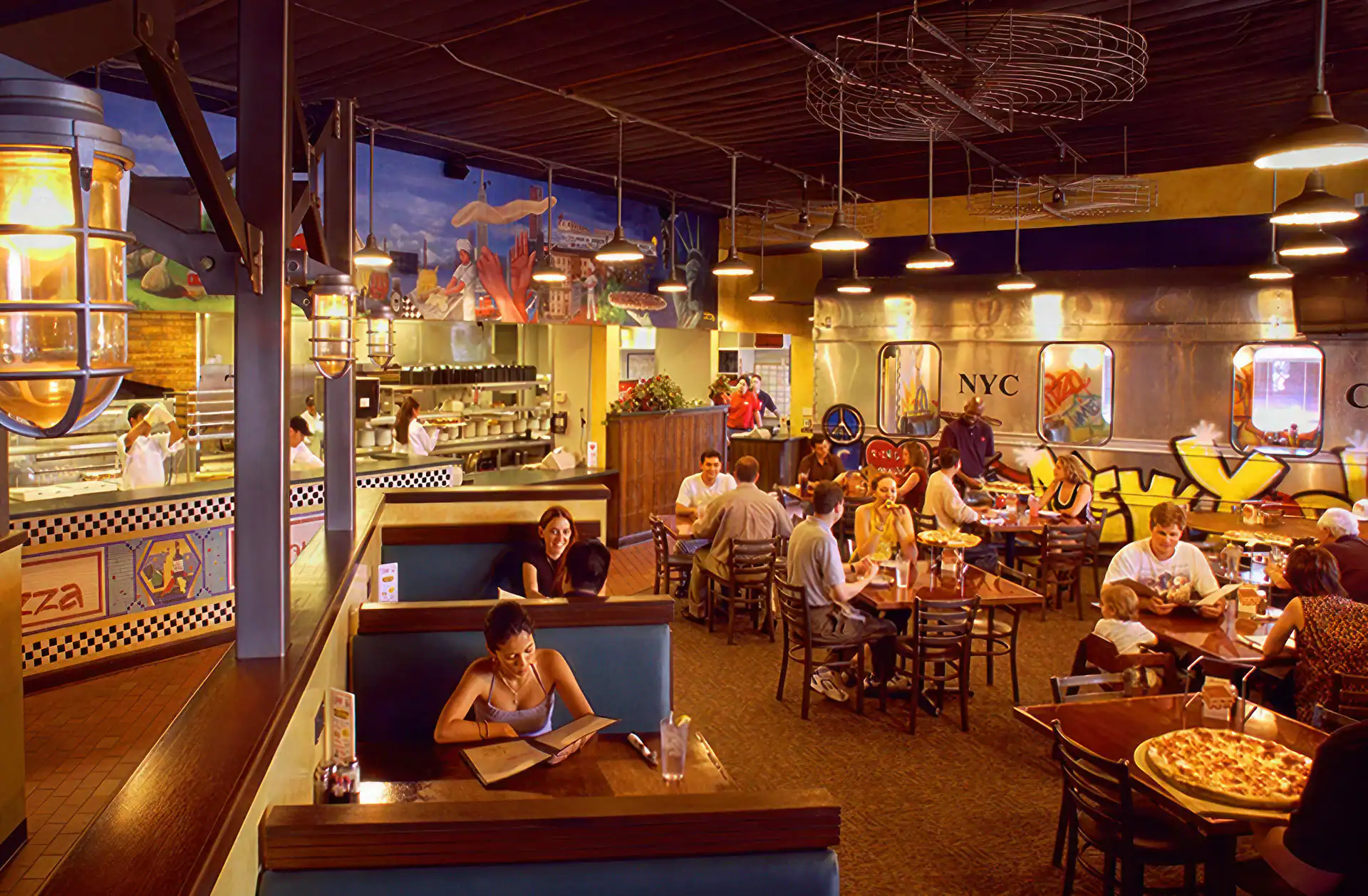 A lively pizza restaurant in TX with colorful murals, groups dining, some reading menus, and staff by an open kitchen. Booths and tables fill the space&mdash;perfect for capturing moments with top photography services Harris County offers.