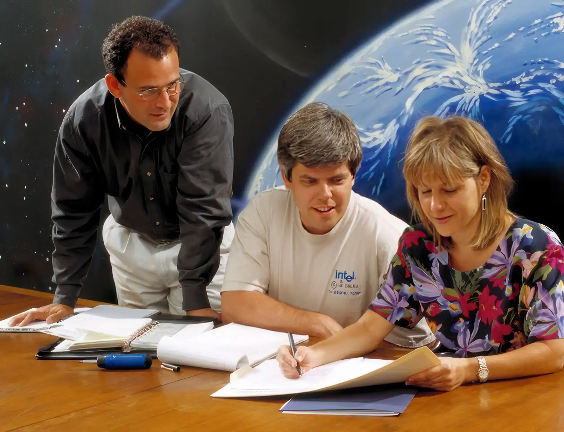 Three people sit at a table covered with papers in a TX office, reviewing documents. A man stands nearby as a woman writes. A large image of Earth from space, perfect for photography services Harris County, hangs on the wall behind them.