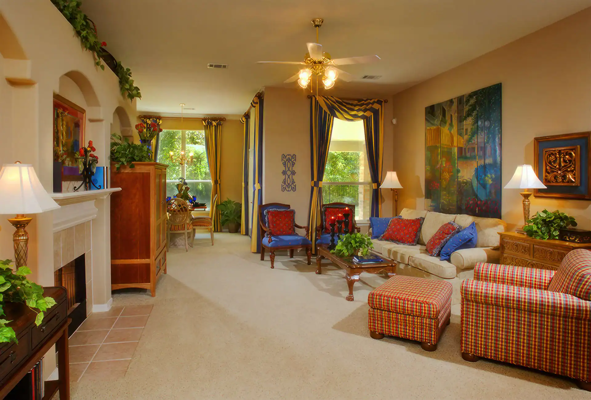 A spacious, brightly lit living room in TX with beige carpet, a cream sofa with colorful pillows, plaid ottomans, blue armchairs, plants, lamps, artwork, and large windows—perfect for photography services in Harris County.