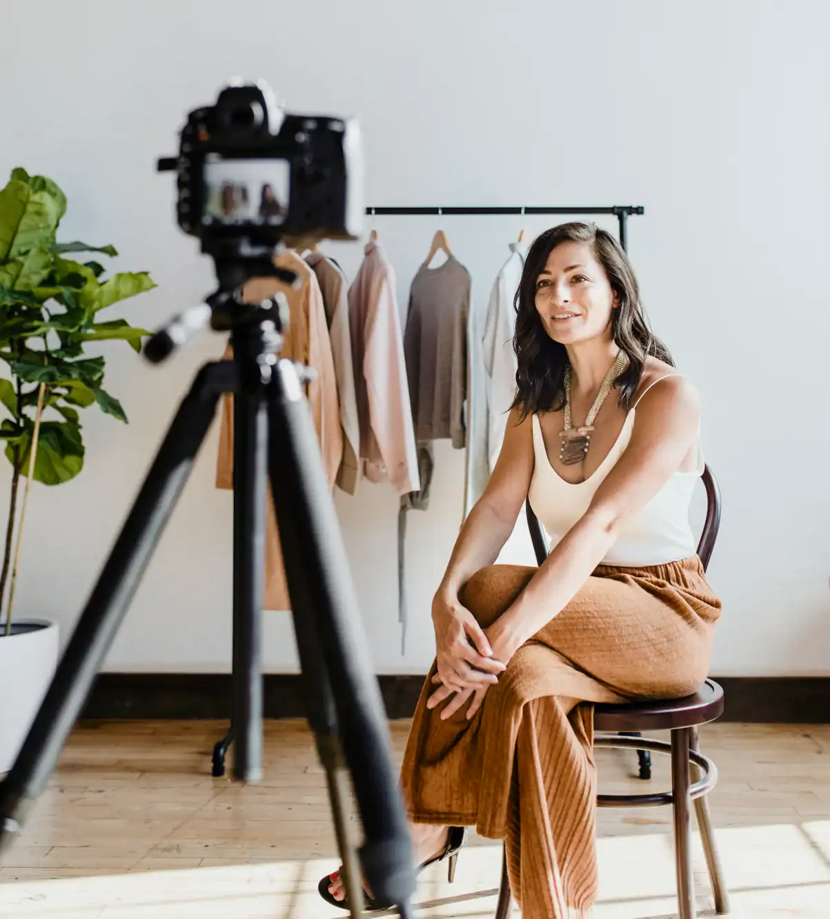 A woman sits on a chair in front of a camera on a tripod, smiling. Behind her, a clothing rack displays neutral-toned garments. In this bright, minimalist TX studio, the photography service Harris County trusts captures authentic moments.