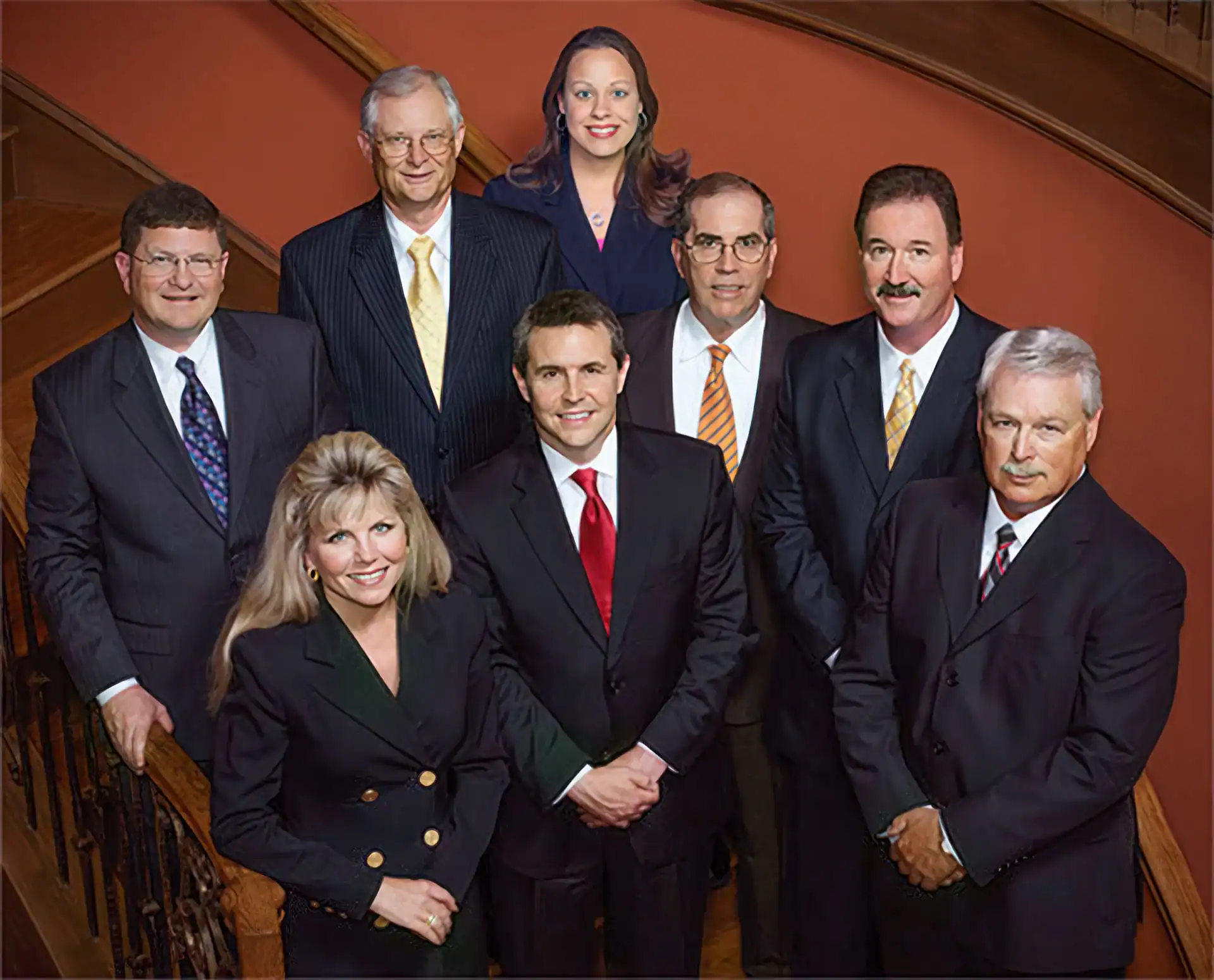 Seven men and one woman in business attire pose on a staircase, smiling at the camera. The formal setting, with wooden railings and steps, highlights the professionalism of photography services in Harris County, TX.