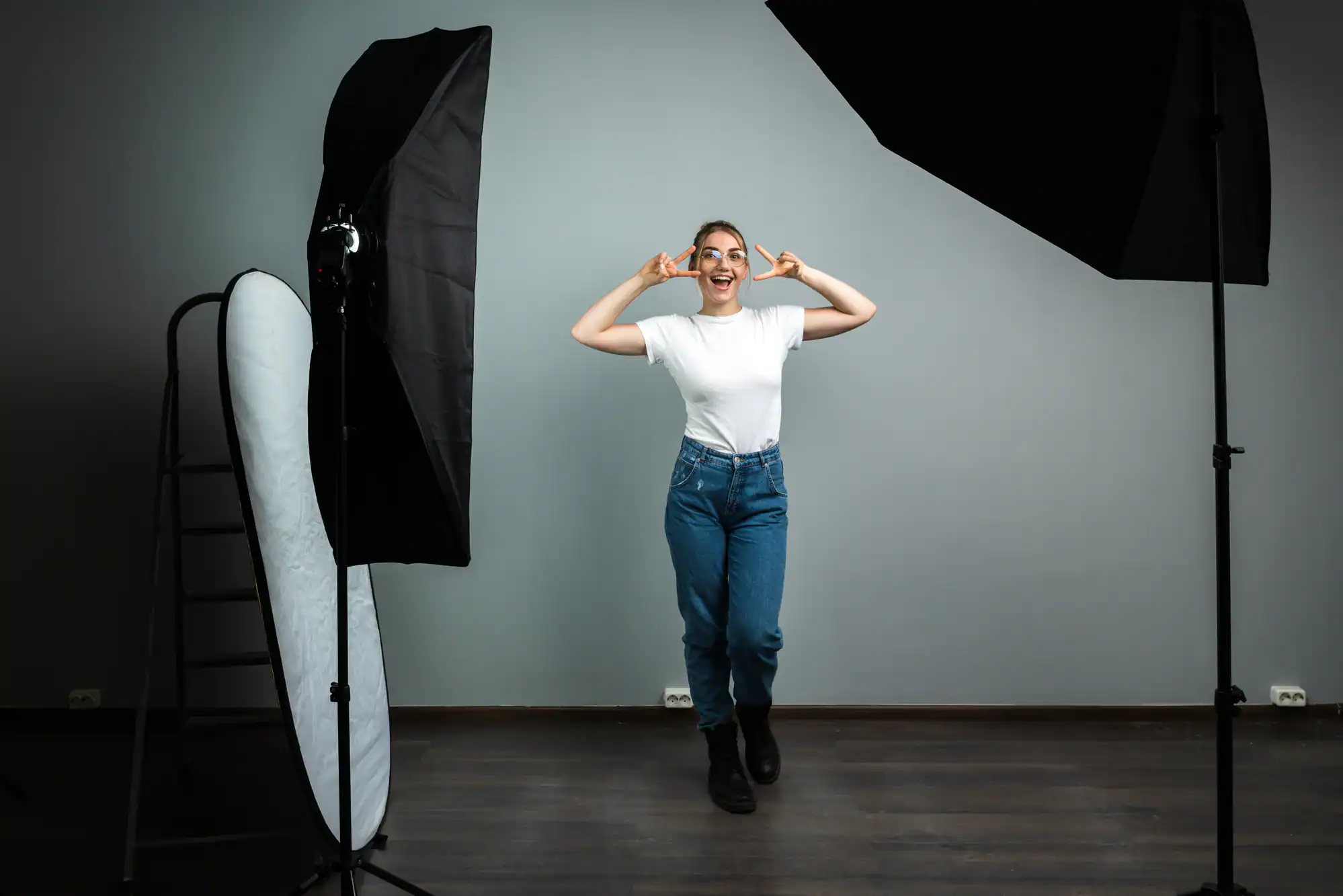 A person in a white t-shirt and jeans poses playfully in a photography studio, making a peace sign with both hands. Studio lights and a reflector surround them—a lively moment from a photography service in Harris County, TX.