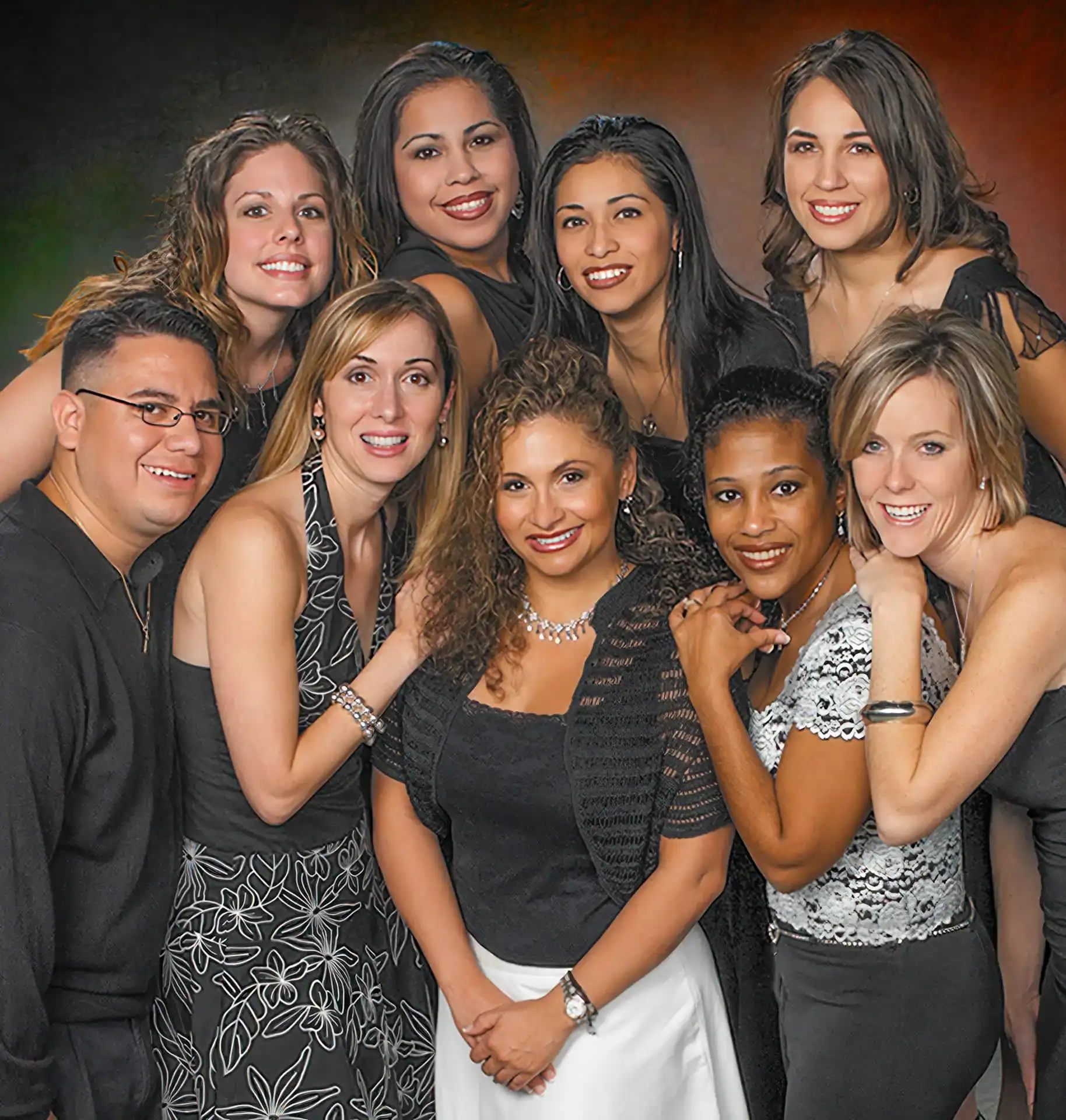 A group of nine adults, eight women and one man, dressed in formal black and white attire, smiling and posing closely together against a dark, blurred studio backdrop&mdash;showcasing professional photography services in Harris County, TX.