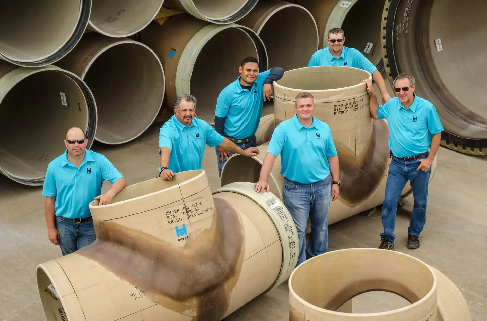 Six men wearing matching light blue shirts stand and pose among large industrial pipes or fittings, smiling at the camera outdoors—a great example of group photography services in Harris County, TX.