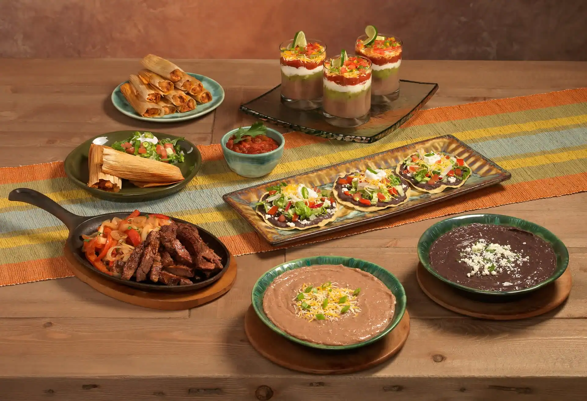 A wooden table set with Mexican dishes like tamales, flautas, beef strips with peppers, tostadas, and more on colorful placemats, perfect for TX gatherings or highlighting photography services in Harris County.