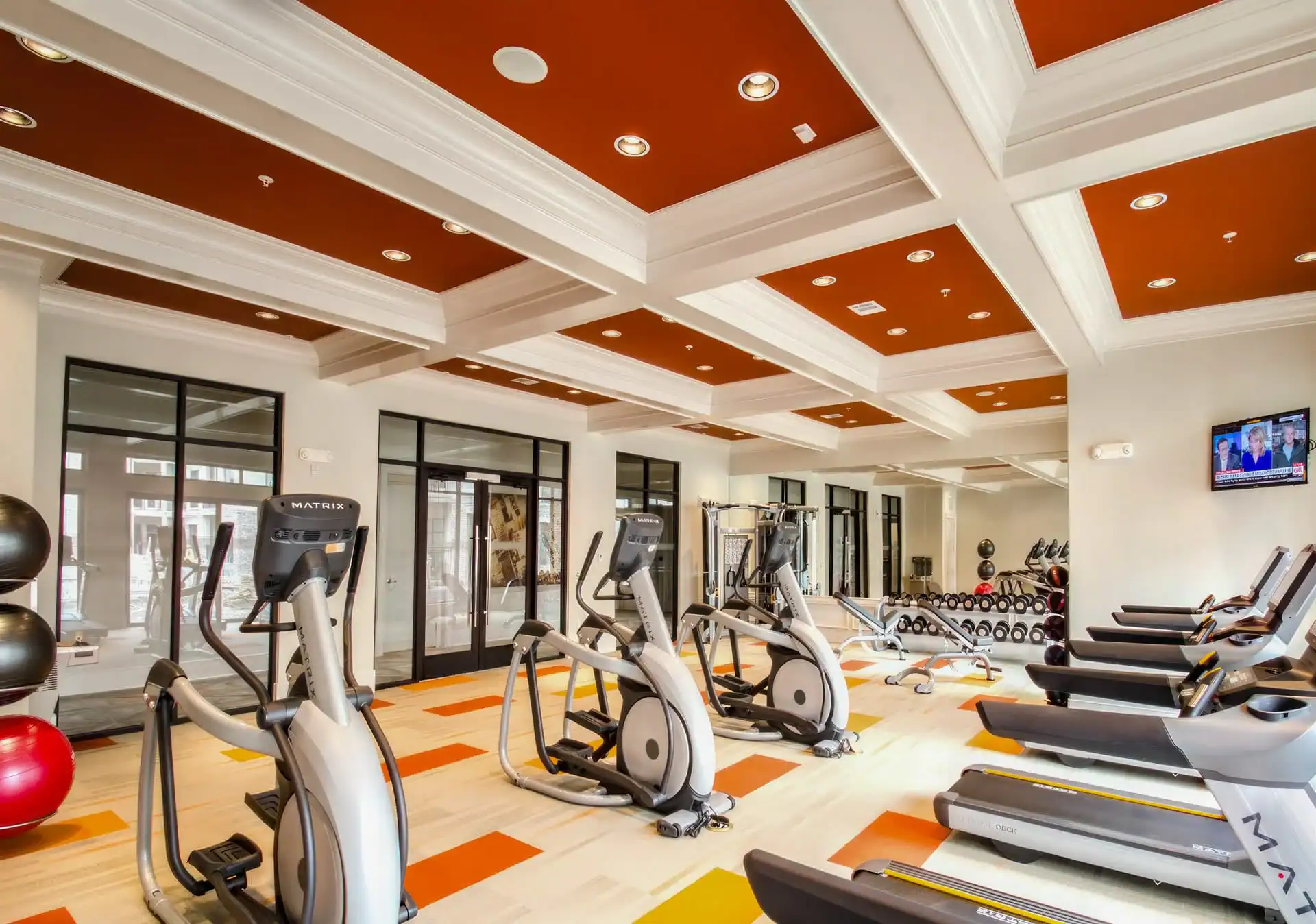A modern gym with treadmills, ellipticals, exercise balls, and free weights on colorful orange, yellow, and white flooring. Large windows and mirror walls reflect the bright interior&mdash;ideal for photography services in Harris County, TX.