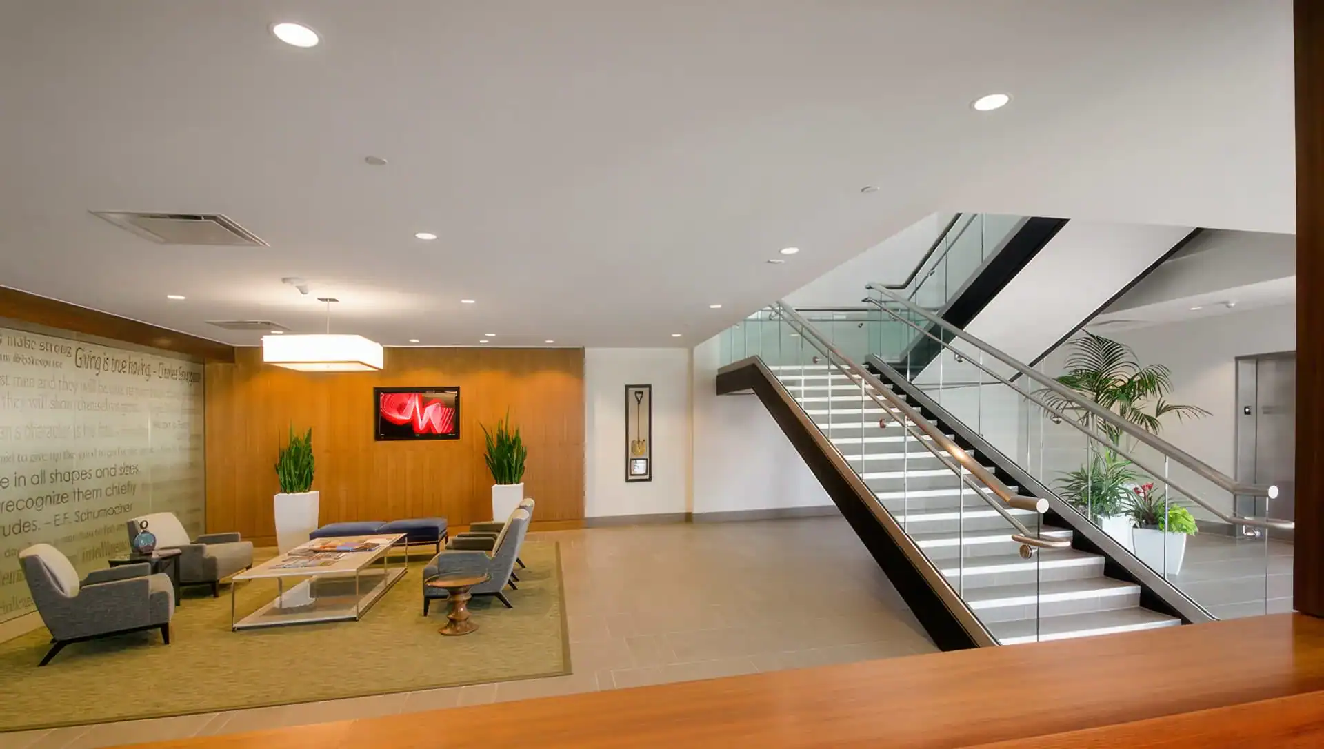 Modern office lobby with seating area, four gray chairs, a glass coffee table, TV on a wooden accent wall, plants, and a wide staircase with glass railing—perfect for showcasing with professional photography services in Harris County, TX.