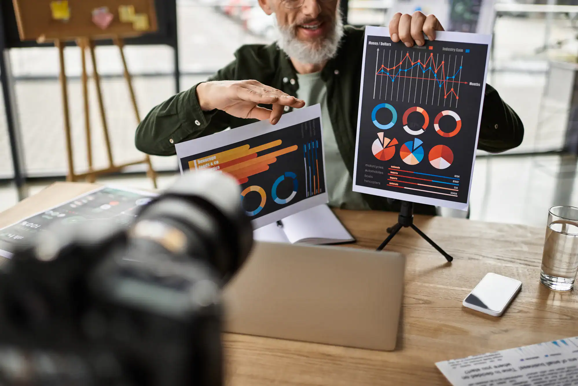 A person in TX presenting colorful charts and graphs on printed papers during a meeting, with a camera recording—ideal for a photography service Harris County—with a laptop, notebook, glass of water, and smartphone on the wooden table.