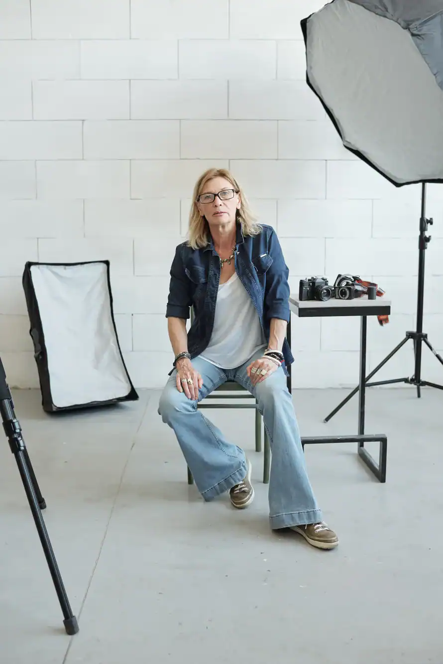 A woman with blond hair and glasses sits on a stool in a TX photography studio, surrounded by lighting equipment, cameras on a table, and wearing a denim jacket, flared jeans, and sneakers—ready to offer her photography service in Harris County.