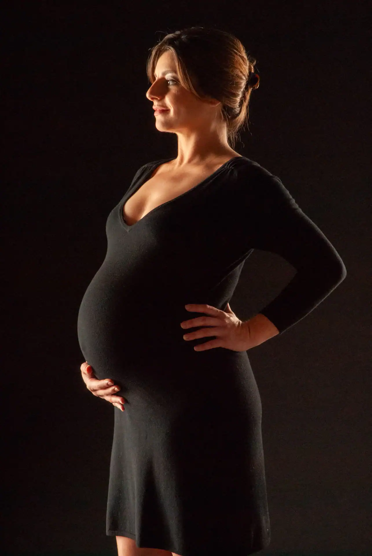 A pregnant woman in a black dress stands in profile against a dark background, one hand on her hip and the other gently resting on her belly, softly lit from the side&mdash;perfect for photography services in Harris County, TX.