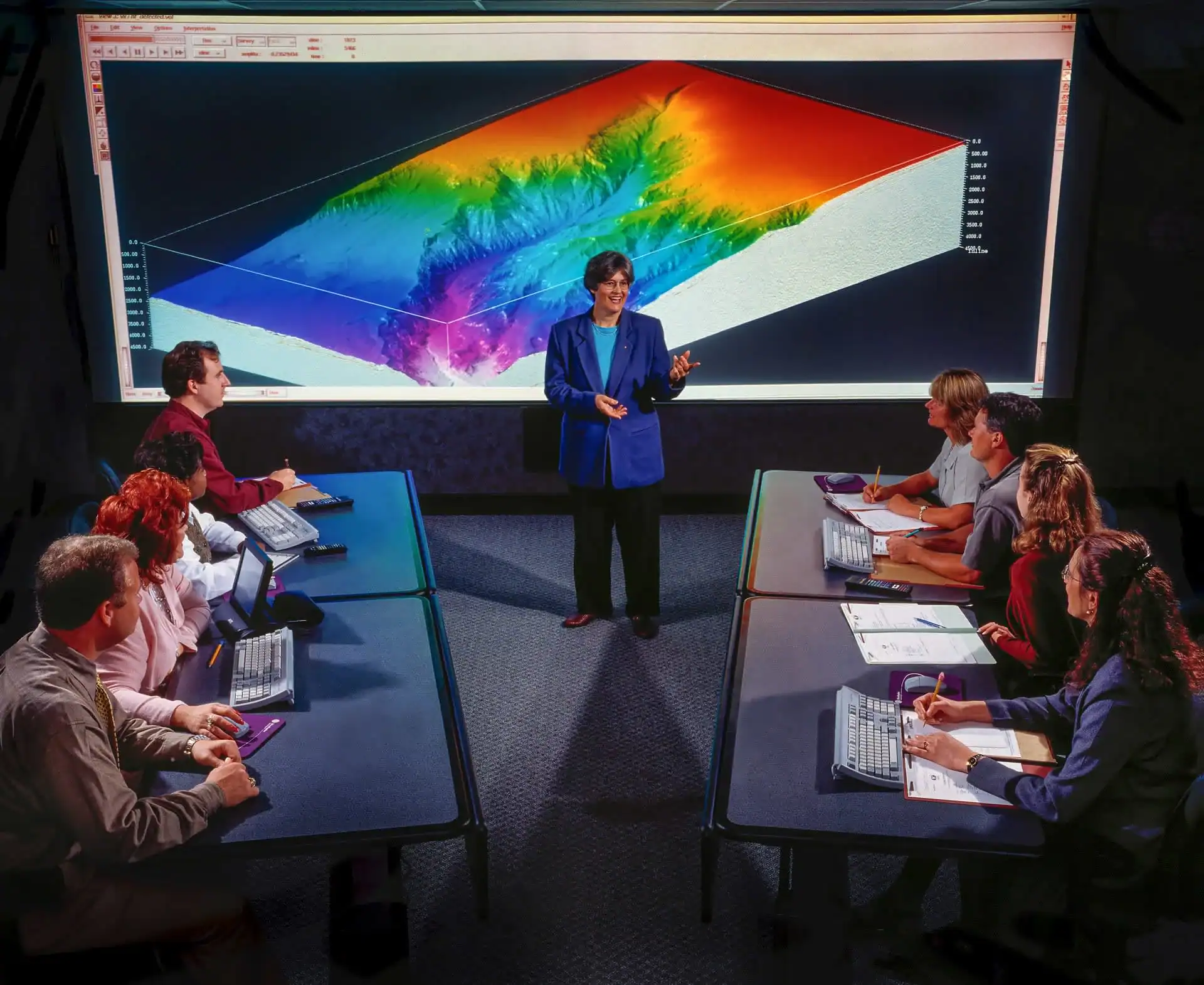 A person stands presenting in front of a large 3D topographical map displayed on a screen, while eight people at desks with computers listen and take notes—ideal for showcasing photography services in Harris County, TX.