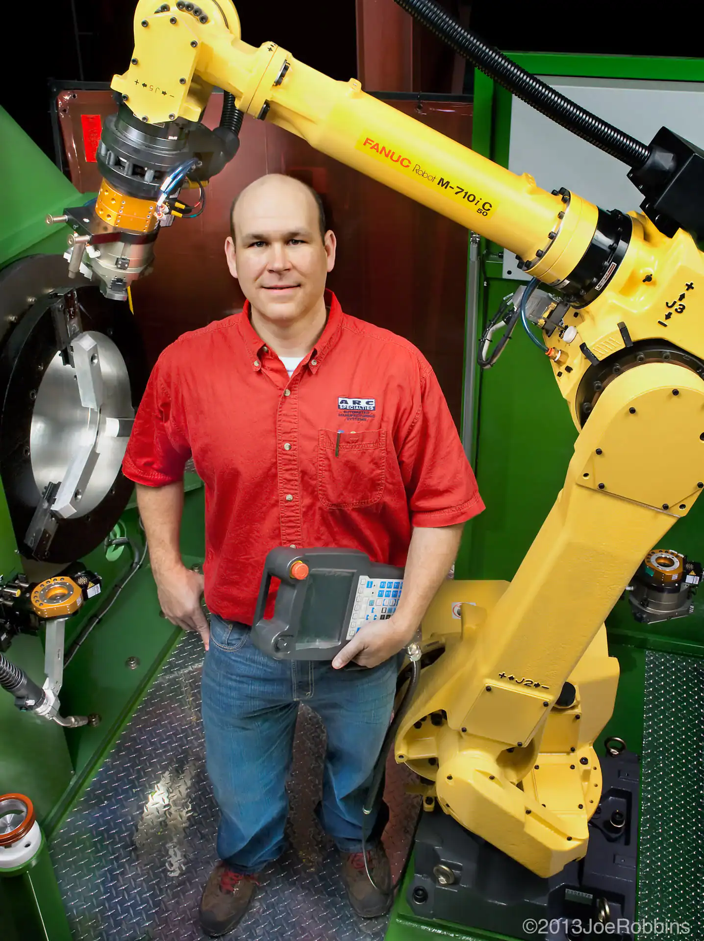 In TX, a man in a red shirt stands next to a large yellow industrial robot arm, holding a control panel inside a modern manufacturing facility. The environment features green and silver machinery&mdash;ideal for photography services Harris County.