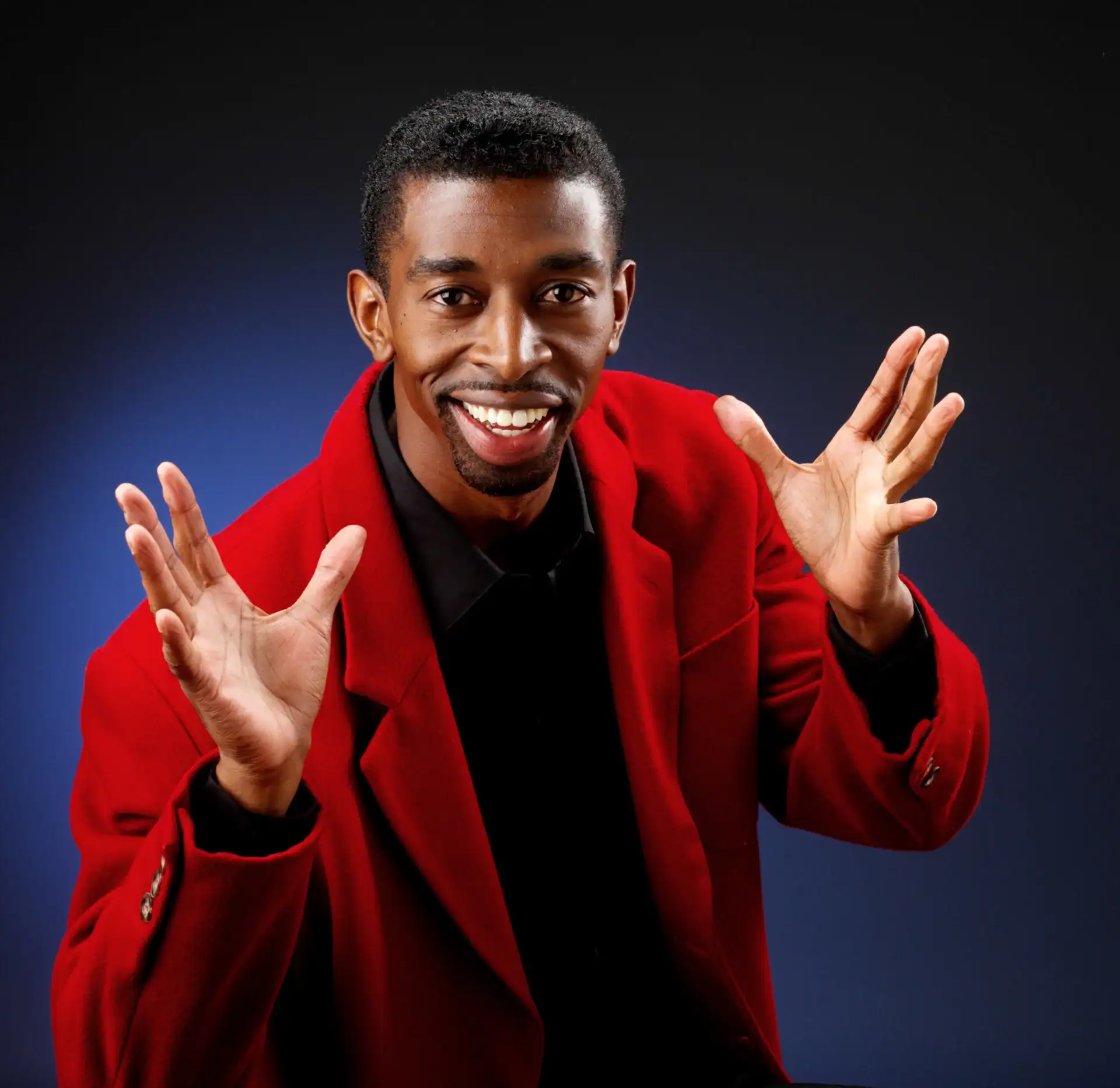 A smiling man in a bright red jacket and black shirt gestures energetically with both hands raised, set against a dark blue and black gradient background&mdash;perfect for showcasing TX photography services Harris County.