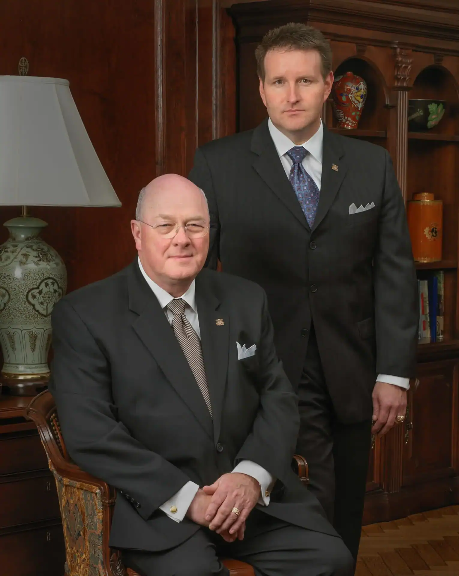 Two men in suits pose in an elegant TX office; one sits while the other stands behind him. A lamp, bookshelf, and wooden cabinet with decorative items set the scene&mdash;ideal for showcasing professional photography services Harris County.