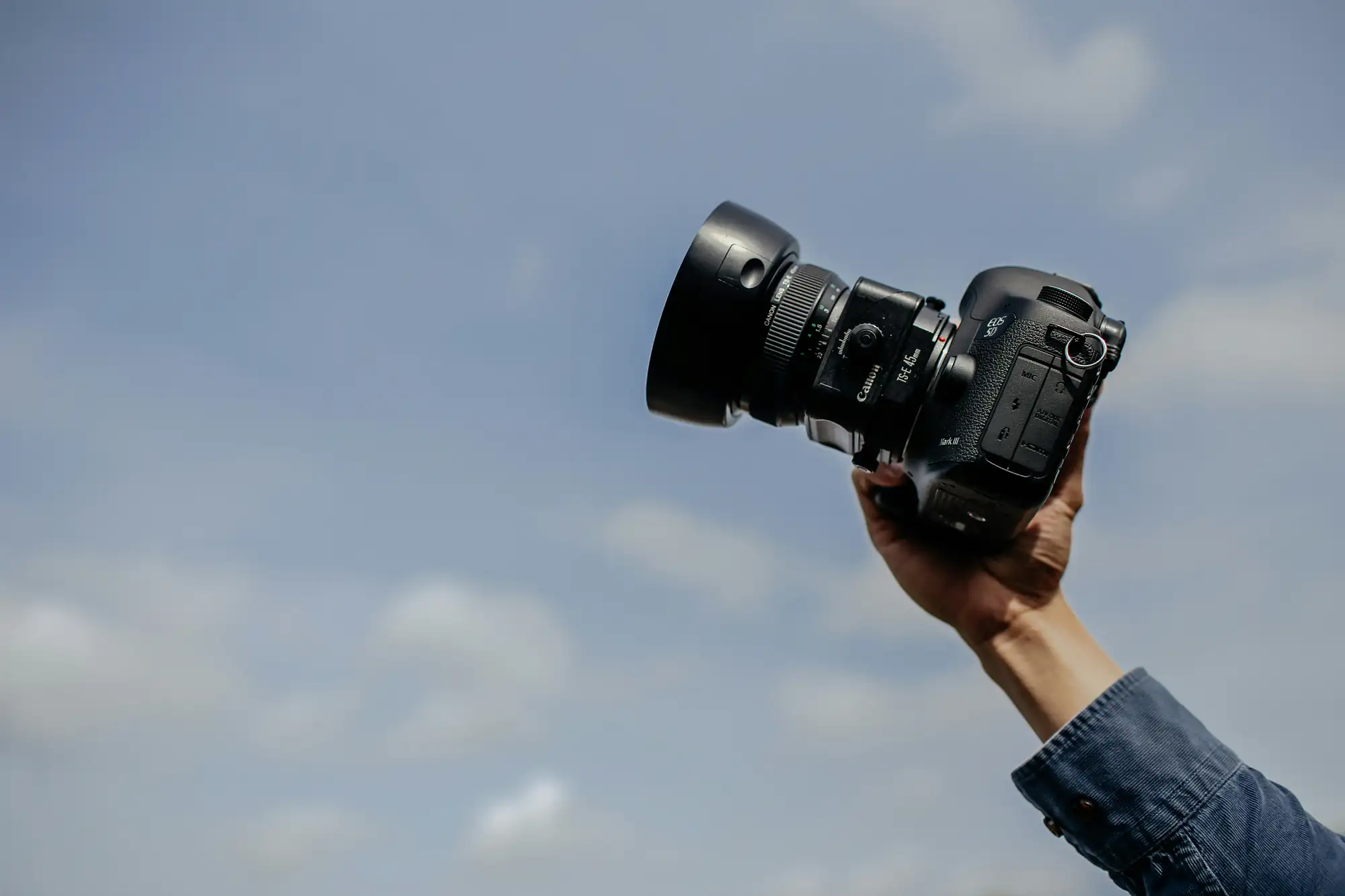 A hand holds a DSLR camera against a bright blue sky with scattered clouds, capturing photography moments.
