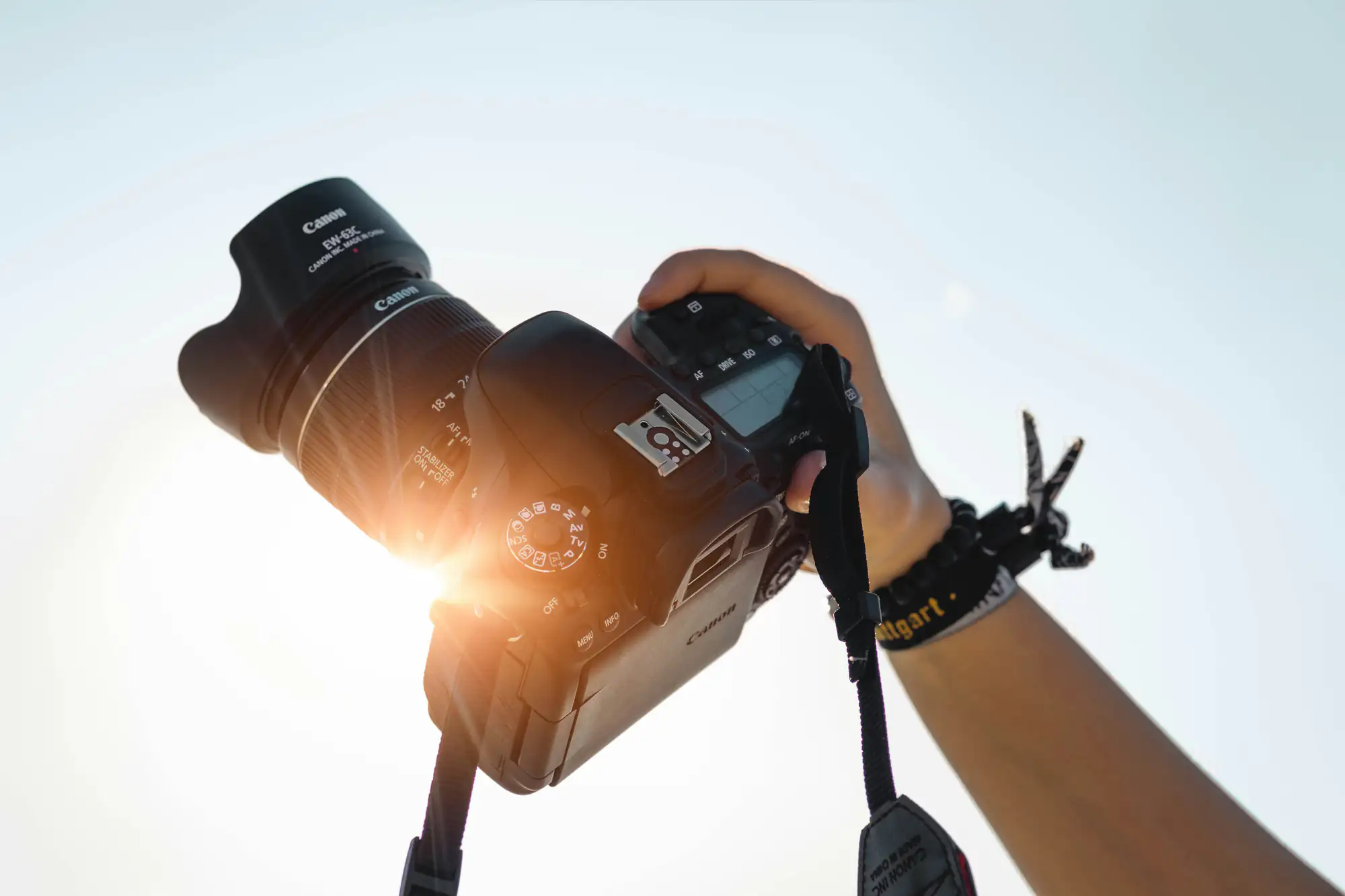 A hand holds a DSLR camera toward the sky as sunlight shines through the lens in this photography image.